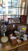 Photo showing a collection of miscellaneous decorative items including a cream-colored lamp with glass globe, framed floral print, woven baskets, a doll figure, jars, various small items, and a box.