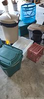 Photo showing the two large plastic outdoor garbage cans, multiple storage totes in green and white, blue mesh organizer, and red crate.