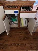 Wood cabinet with two drawers, one drawer contains silverware sets including spoons, forks, and knives with some plastic utensils visible; lower shelves have various plastic containers and lids stacked together