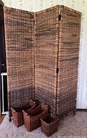 Full view of three-panel woven rattan dressing room screen with four assorted baskets in front on carpeted floor against floral wallpaper.