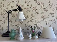Wide view of bronze desk lamp with white fabric shade, assorted small pottery pieces and décor on surface against floral wallpaper background.