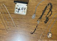 Photo showing sterling silver cameo brooch, Creed Jewelry rosary with black beads, four chain bracelets, and assorted religious charms laid out on a wooden surface.