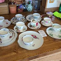Overall assortment of ten teacups set on saucers, along with some snack plates showing floral designs and gold trim, displayed on a wooden table.