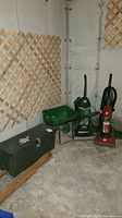 A corner photo showing two upright vacuum cleaners, a green wheelbarrow, and a black tool chest on the concrete floor against a lattice wall.
