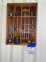 Wooden display case containing multiple vintage miniature metal objects including pots, pitchers, spoons, and figurines arranged in individual compartments.