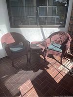 Two reddish-brown wicker chairs and one matching wicker table on a brick floor near a window, lit by sunlight.