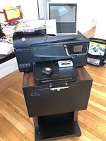 HP OfficeJet 6700 printer with touchscreen on top of rolling filing cabinet, Amazon Echo Show screen next to it, power cables visible