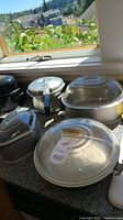 Photo shows multiple cookware pieces on countertop including Guardian Service pressure cooker, glass lidded pots, roasting pans, and metal trays under natural light.