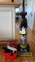Full view of Bissell upright vacuum cleaner with red container of extra parts beside it on floor.
