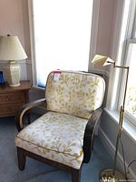 Wood chair with curved wooden arms and cream cushions with yellow floral pattern positioned by window, next to side table lamp and brass floor lamp.