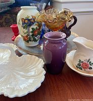 White ceramic vase with blue bird and flowers, amber glass vase with ruffled edge, small purple vase, white pitcher with rose, cream shell-shaped plate, red candlestick holder on dark wooden surface