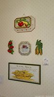 Wall display showing vegetable-themed ceramic plaques and framed kitchen art, with a cuckoo clock above