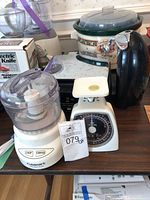 Lot contents on table showing Rival slow cooker, Black and Decker waffle maker, Hamilton Beach can opener box, Cuisinart food processor and mechanical kitchen scale.