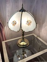Glass and brass table lamp lit on glass table with floral motif on shade, 23 inches tall