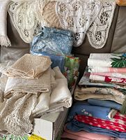 Stacked cream-colored crochet doilies and linens with assorted colorful folded fabric linens and tablecloths behind on couch.