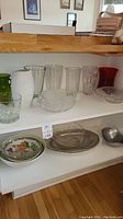Shelves displaying assorted glass and ceramic bowls, plates, and vases including clear glass, green and red glass, and white ceramic items.