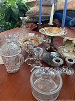 Overview of silver-toned serving pieces, candlesticks with candles, cut glass containers, and small decorative items arranged on a surface.