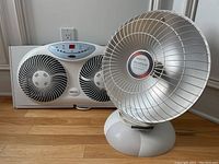 Photo showing both the white double blade window fan and the Presto HeatDish heater unit placed on a wooden surface against a wall.