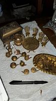 Overview of brass miniatures arranged on cloth with pen for scale, showing rectangular box, miniature chairs around a round table, brass pitchers, candle holders, and small wine glasses.