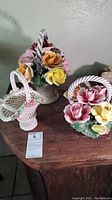 Two Capodimonte porcelain flower baskets displayed on a wooden surface; both showing signs of damage.