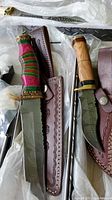 Two collector knives with patterned blades, multicolored and natural wood handles, and leather sheaths.