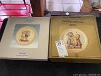 Photo of two large Hummel plates in their original boxes with visible age on boxes, showing design images on the front.