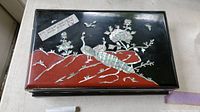 View of rectangular black lacquer box with mother of pearl inlay depicting peacocks and flowers on red rocky base