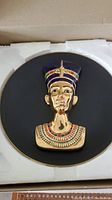 Collector plate featuring a gold-tone and enamel three-dimensional bust of Nefertiti, set on a black background and encased in protective foam in a box.