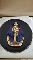 Porcelain collector plate, black background with raised painted depiction of King Osiris in gold, blue, red, and green details.