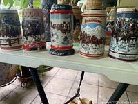 Five Budweiser branded collectible beer steins displayed on a table, decorated with winter scenes and horse-drawn carriages.