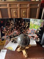 Photo of lacquer folding table screen behind dark marble accent ball on gold-toned X stand on wooden table.