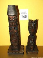 Photo showing two hand carved wooden totems against yellow background, size comparison with auction lot number card visible.