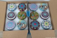 Two open boxes showing 24 round candles in tins with various colorful decorative lids arranged in rows on foam padding.