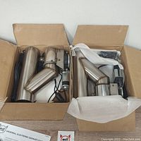 Two boxes, each containing a stainless steel universal electronic exhaust cutout kit with electric actuator and wiring, viewed from above inside packaging