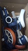 Box filled with disassembled children's tricycle parts including wheels, seats, handles, and frame components in blue, white and orange.
