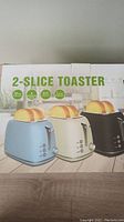 Photo of the box showing three color options for the 2-slice toaster with toasted bread on top.