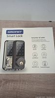 Box front showing Smonet Smart Lock image and key features including key free access, app control, anti-theft alert, auto-lock, battery backup, and touchscreen keypad.