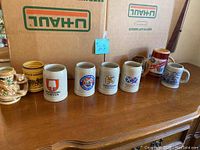 Photo showing nine assorted beer mugs arranged in a row on a wooden surface with cardboard boxes in the background