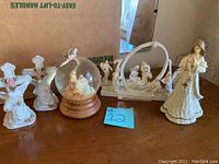 Photo of eight Christmas nativity and angel-themed figurines, including a snow globe and a white arch nativity scene, displayed on a wooden surface.