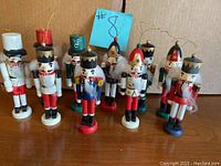 Photo of 10 mini nutcracker soldier figure ornaments, all standing upright with hanging loops on top, placed on a wooden surface with a blue paper label numbered 8 in the background.
