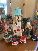 Photo showing various Christmas decorative figurines including a white birdhouse with pinecones and rustic tools, two caroler figurines in painted clothing holding hymn books, two ceramic snowman tealight holders, and a lamppost wrapped with pine garland and a red plaid bow. Items show signs of dust.