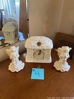 Photo of three white ceramic candle holders, including a central house-shaped holder with star cut-out and two angel holders on either side, displayed on a wooden surface.