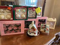Photo shows three unopened boxes of hand-decorated glass Christmas ornaments (two white and one light pink), two unopened boxes of 10 light rose string lights, a ceramic musical Christmas village snow globe with children, and a ceramic Christmas music box with snowman and child figurines.