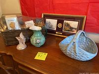 Framed Thomas Kinkade prints, green Fenton pitcher, pink iridescent Fenton butterfly figurine, boxed M.J. Hummel ceramic basket in a wicker gift basket, and light blue ceramic basket displayed on wooden table with red background.