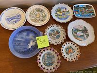 A photo showing nine collectors plates with various scenic, map, and floral designs in different shapes including round, scalloped, pierced edges, and rectangular.