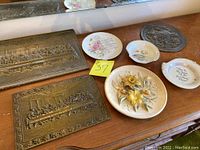 View of two Last Supper embossed metal plaques and five decorative plates with floral and religious script designs on wooden surface.