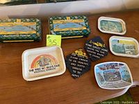 Photo showing two McEwan's Guest Edinburgh collector tins, four trivets with various designs including a black trivet with a quote, and two souvenir trays, one from the 1982 World's Fair in Knoxville, Tennessee and a multiview scenic design tray.