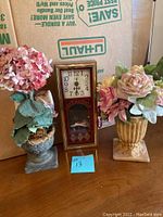 Photo showing two artificial flower arrangements in decorative vases on either side of a vintage-style clock.