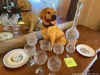 Photo showing wine decanter, six wine glasses, dog figurine, and two decorative plates on wooden surface with mirror background