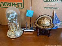 Anniversary clock under glass dome, two sailboat models, and vintage globe on wooden tripod stand shown together on cardboard background.